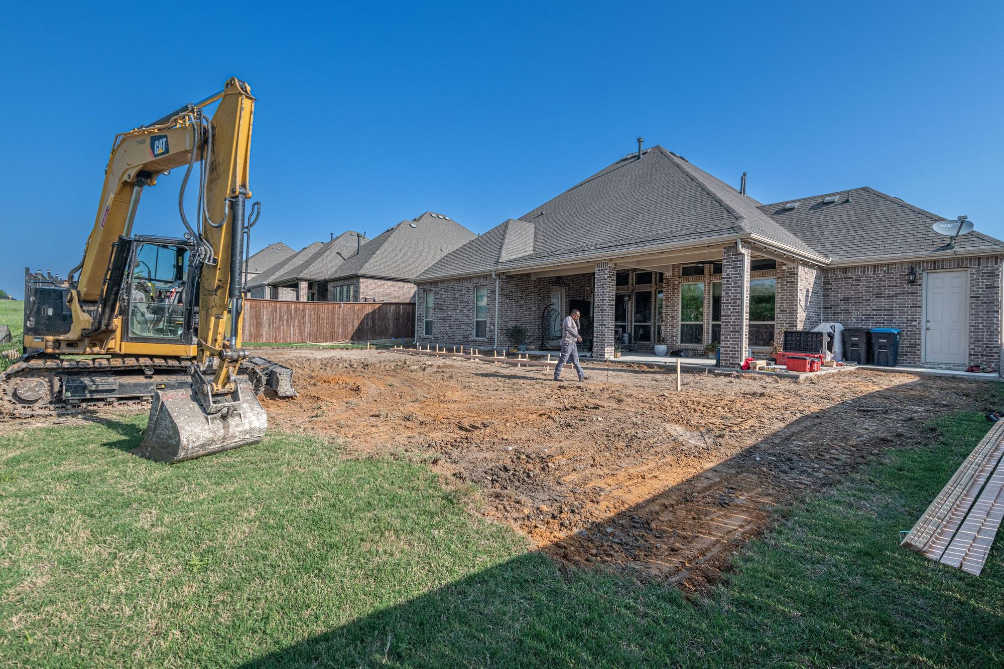 Back yard under construction: bare graded clay and an excavator behind a brick house