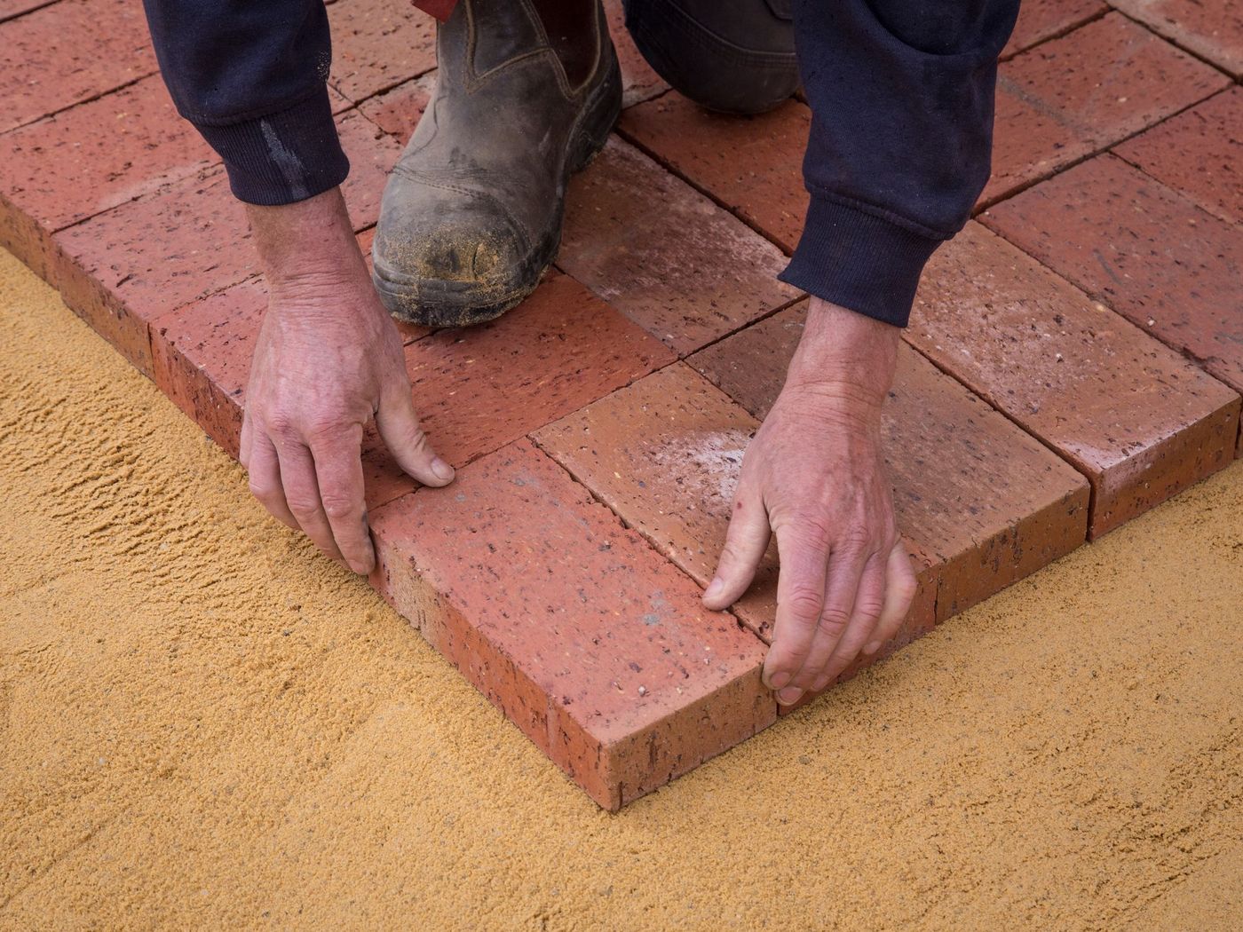 Hands laying clay brick pavers on a sand bed