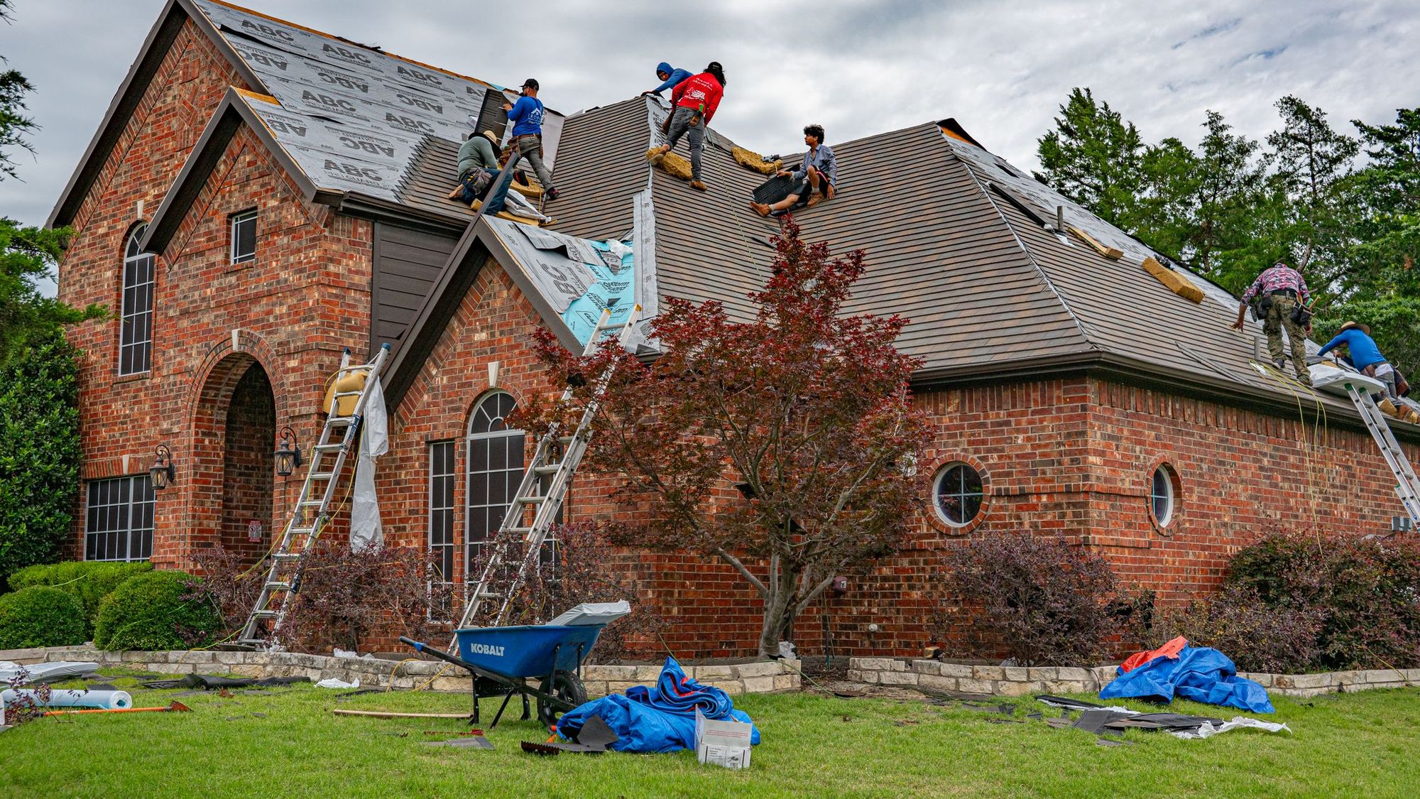 Roofing crew tearing off an old roof on a brick two-story, ladders up and the beds covered